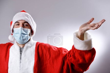 Photo for Young santa claus wearing protective face mask spreads his hands on gray background - Royalty Free Image