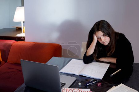 Foto de Chica joven vestida de negro hace su tarea en la mesa de la sala de estar con el ordenador, mirando muy aburrido - Imagen libre de derechos