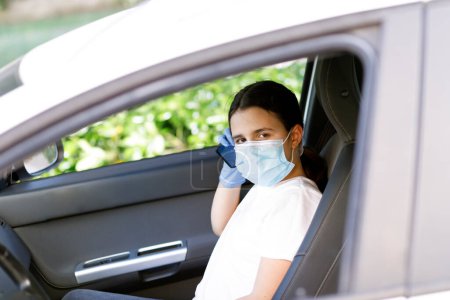 Téléchargez les photos : Jeune fille avec masque et gants regarde par la fenêtre de l'intérieur d'une voiture - en image libre de droit