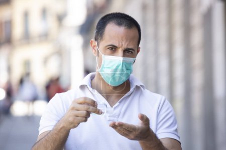 Photo for Dark-haired man with white polo and facial mask disinfects his hands with an antibacterial gel stopped in a sidewalk - Royalty Free Image