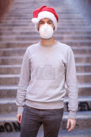 Photo for Man in santa hat, gray shirt and ffp2 mask looks sad isolated on stairs - Royalty Free Image