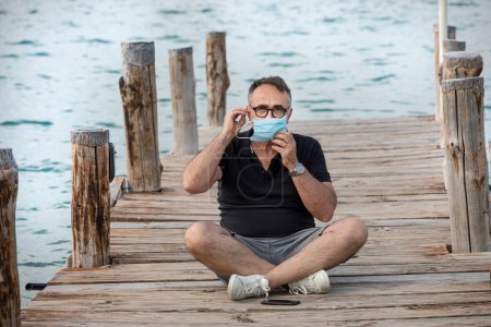 Photo for Man on vacation sitting on a dock in a port puts on a surgical mask - Royalty Free Image