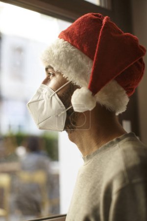 Téléchargez les photos : Garçon avec chapeau de Père Noël et masque de protection semble triste à l'extérieur de la maison à travers une vitre - en image libre de droit