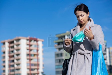 Foto de Chica morena con abrigo, bolsa de compras, se pone su mascarilla, en los edificios de fondo de un barrio de la ciudad y el cielo azul - Imagen libre de derechos