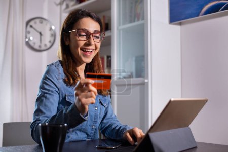 Téléchargez les photos : Brunette fille avec chemise en denim assis dans la table de salon achète en ligne à partir de sa tablette grâce à sa carte de crédit - en image libre de droit