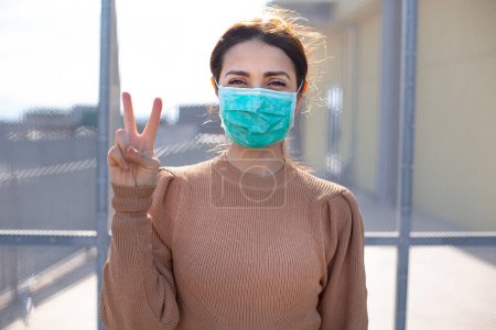 Photo for Brunette girl with orange sweater and facial mask makes the Victory "v" sign with her hand, isolated on wire mesh. - Royalty Free Image