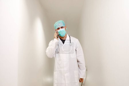 Téléchargez les photos : Médecin avec masque et capuchon dans une salle d'hôpital parle sur un téléphone portable - en image libre de droit