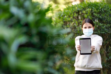 Foto de Chica joven con máscara facial utiliza su tableta en un parque - Imagen libre de derechos