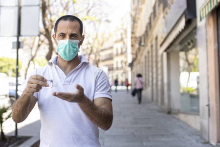 Foto de Hombre de pelo oscuro con polo blanco y máscara facial desinfecta sus manos con un gel antibacteriano detenido en una acera - Imagen libre de derechos