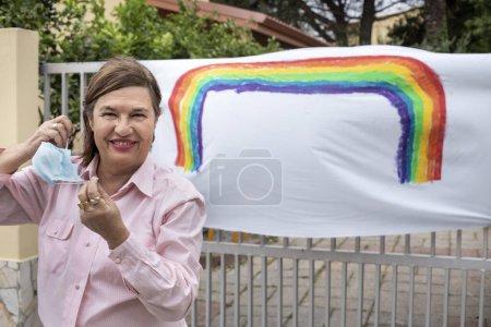 Téléchargez les photos : Femme âgée occasionnel habillé devant un dessin arc-en-ciel sur une porte à l'extérieur de leur maison - en image libre de droit