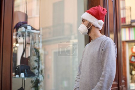 Photo for Young man in medical mask with christmas hat looking at a shop window - Royalty Free Image