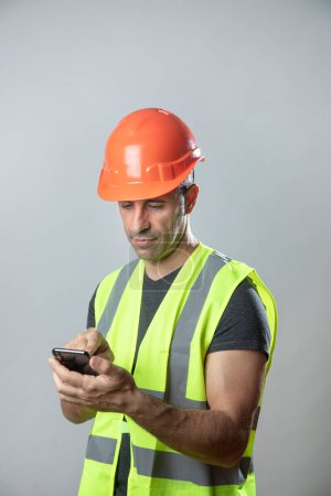 Photo for Worker with orange protective helmet and yellow vest sends an sms from his cell phone, isolated on gray background - Royalty Free Image