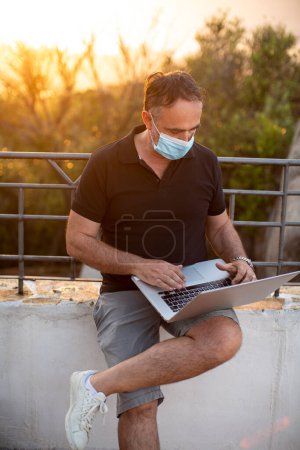 Téléchargez les photos : Homme en vacances habillé occasionnel travaillant sur ordinateur portable protégé par un masque facial - en image libre de droit