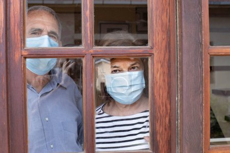 Photo for Elderly couple wearing face masks watch the world through their home window - Royalty Free Image