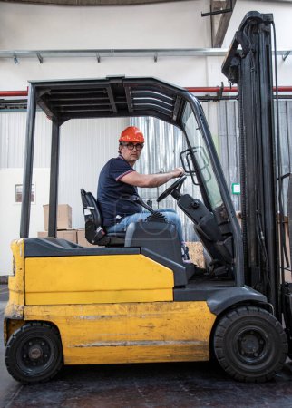 Téléchargez les photos : Travailleur avec casque de protection et lunettes noires conduit un chariot élévateur à fourche à l'intérieur d'un entrepôt - en image libre de droit