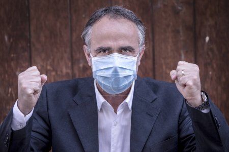 Photo for Man dressed in suit with protective face mask clenches his fists in victory - isolated on wooden background - Royalty Free Image
