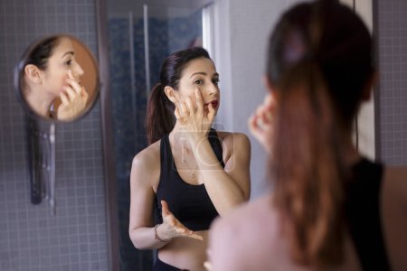 Téléchargez les photos : Belle fille blanche aux cheveux noirs porte du maquillage dans le miroir de salle de bain de sa maison - en image libre de droit