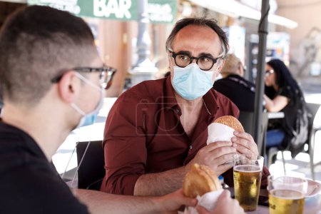 Photo for Father and son wearing casual clothes with masks eating burgers while sitting at the bar - Royalty Free Image