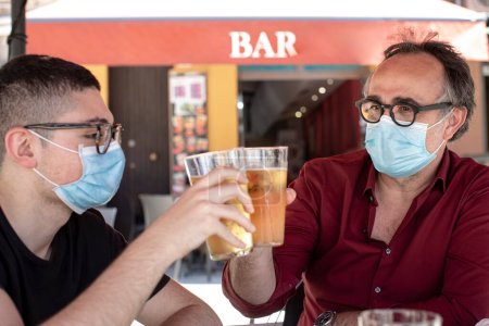 Photo for Young man and an adult sitting at a table with a face masks toast by joining their glasses full of beer - Royalty Free Image
