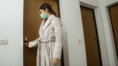Téléchargez les photos : Brunette fille avec chemise violette sort de son appartement portant un masque protecteur - en image libre de droit