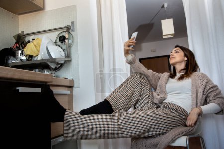 Téléchargez les photos : Casual habillé brunette fille assise dans la cuisine sur une chaise prendre un selfie avec son smartphone - en image libre de droit