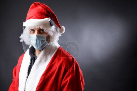 Téléchargez les photos : Père Noël avec masque chirurgical, bouclier protecteur en plexiglas et chapeau de Père Noël semble grave, isolé sur fond noir - en image libre de droit