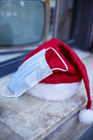 Photo for Santa Claus hat with a surgical mask nearby and in the background behind a shop window there is a vintage television - Royalty Free Image