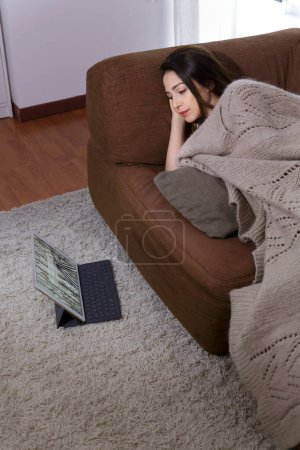 Foto de Mujer joven mira su tableta colocada en la alfombra mientras ella está acostada en el sofá - Imagen libre de derechos