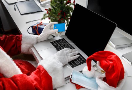 Foto de Detalle de la mano de Santa Claus sosteniendo una tarjeta de crédito listo para realizar un pedido en Internet sentado en un escritorio con un ordenador portátil - Imagen libre de derechos