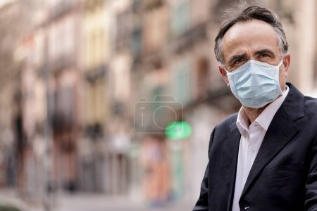 Photo for Portrait of middle-aged man in jacket with protective face mask, standing on a sidewalk with public greenery behind - Royalty Free Image