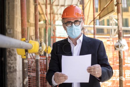 Photo for Engineer, under the scaffolding of a construction site he wears a protective helmet and a surgical mask - Royalty Free Image