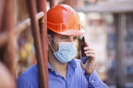 Téléchargez les photos : Ingénieur se tient près d'un chantier de construction de la ville portant un casque de protection et un masque chirurgical lors de l'utilisation de son téléphone portable - en image libre de droit