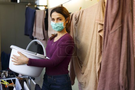 Foto de Chica morena con camisa morada cuelga la ropa en la lavandería del hogar, inodssando la máscara protectora - Imagen libre de derechos