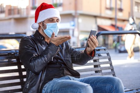 Photo for Man with Santa hat, leather jacket and surgical mask, makes a video call sitting on a bench in the city - Royalty Free Image