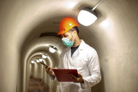 Téléchargez les photos : Ingénieur avec casque de protection, masque chirurgical et manteau blanc tient un dossier rouge à l'intérieur d'un tunnel souterrain - en image libre de droit