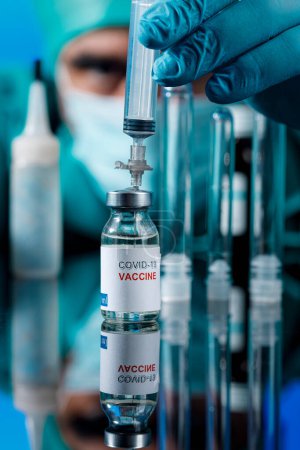 Foto de El médico con gorra médica y guantes de látex recoge la vacuna covid con una jeringa en una mesa con varios viales - Imagen libre de derechos