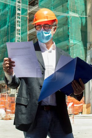 Foto de Ingeniero se encuentra cerca de un sitio de construcción de la ciudad y lleva casco protector y una máscara quirúrgica - Imagen libre de derechos