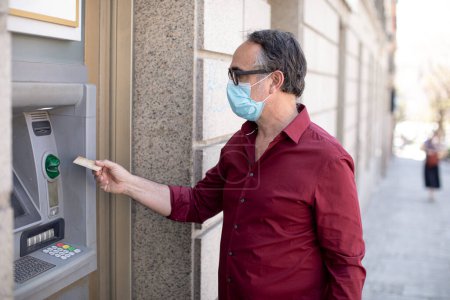 Téléchargez les photos : Casual habillé homme avec masque facial retire de l'argent d'un distributeur automatique dans la rue - en image libre de droit
