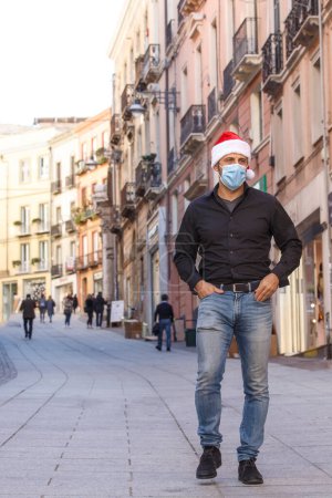 Photo for Man with Santa hat, shirt, jeans and surgical mask, in the city center among the shop windows - Royalty Free Image