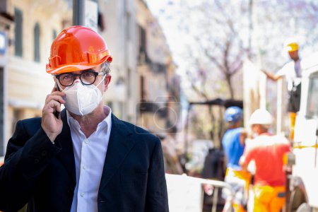 Photo for Engineer stands near a city construction site wearing a protective helmet and a surgical mask while using his cellphone - Royalty Free Image