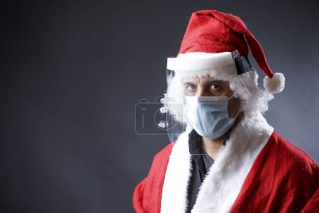 Téléchargez les photos : Père Noël avec masque chirurgical, bouclier protecteur en plexiglas et chapeau de Père Noël semble grave, isolé sur fond noir - en image libre de droit
