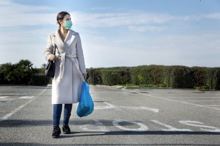 Photo for Girl with coat and facial surgical mask, walks near the supermarket with the shopping bag in hand - Royalty Free Image