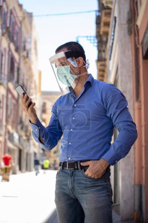 Téléchargez les photos : Homme avec masque habillé décontracté se protège dans la ville tout en parlant sur un téléphone portable - en image libre de droit