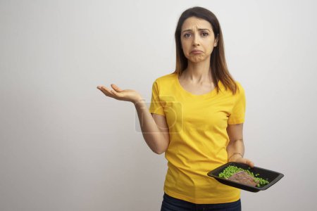 Téléchargez les photos : Jolie fille brune avec t-shirt jaune montre un plat diététique avec air déçu isolé sur fond blanc - en image libre de droit