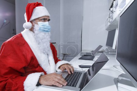 Foto de Santa Claus con máscara quirúrgica, sentado en la oficina, utiliza el ordenador para realizar pedidos a través de Internet - Imagen libre de derechos