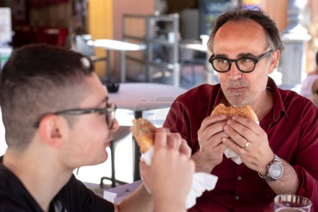 Téléchargez les photos : Père et fils portant des vêtements décontractés mangeant des hamburgers assis au bar - en image libre de droit