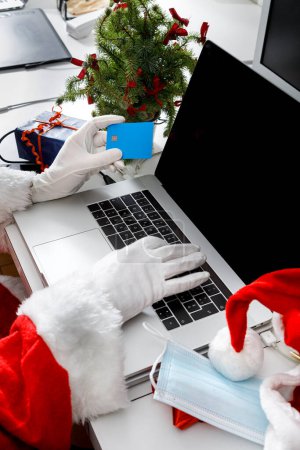Photo for Detail of Santa Claus hand holding a credit card ready to place an order on the internet sitting in a desk with a laptop - Royalty Free Image