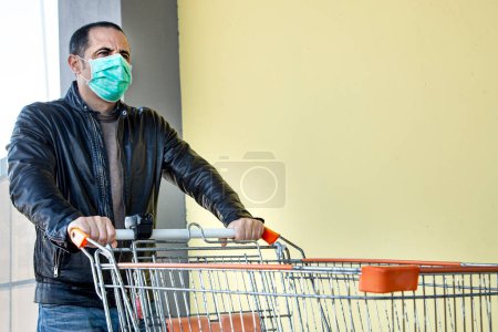 Téléchargez les photos : Casual habillé homme avec veste en cuir et masque protecteur va épicerie au supermarché avec un chariot d'achat, isolé sur un mur jaune - en image libre de droit