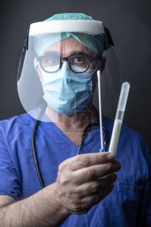 Foto de Médico con protección quirúrgica y bata de laboratorio azul muestra un hisopo faríngeo, aislado sobre fondo negro - Imagen libre de derechos