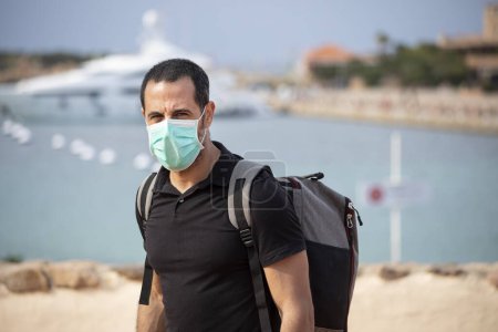 Foto de Hombre moreno en traje de verano y mochila camina por una ciudad balnearia con máscara protectora - Imagen libre de derechos